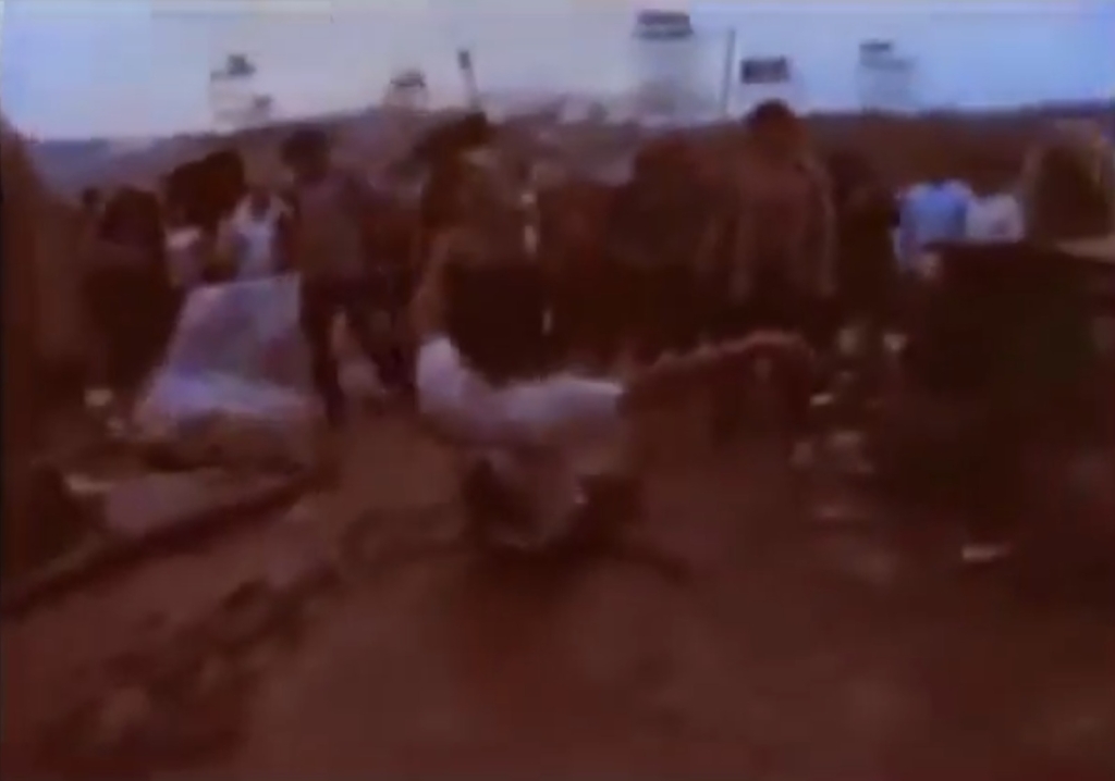 Man sliding in mud, potentially shows bootleg Woodstock print on back of t-shirt