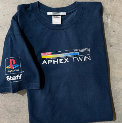 Navy T-shirt laid flat with APHEX TWIN graphic and a loading-bar design, plus a PlayStation Staff logo on the left sleeve.